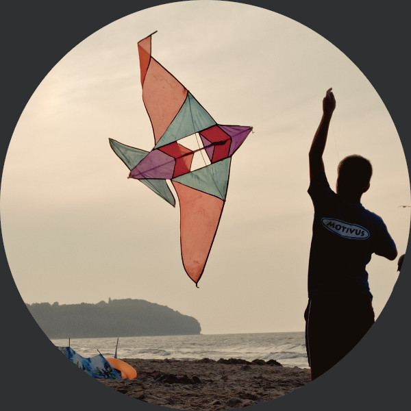 Person am Strand lässt bunten Drachen steigen, Meer und Küstenlinie im Hintergrund bei Sonnenuntergang.