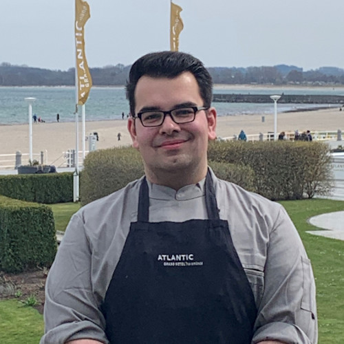 A chef at the ATLANTIC Grand Hotel Travemünde smiles against the backdrop of the Baltic Sea.