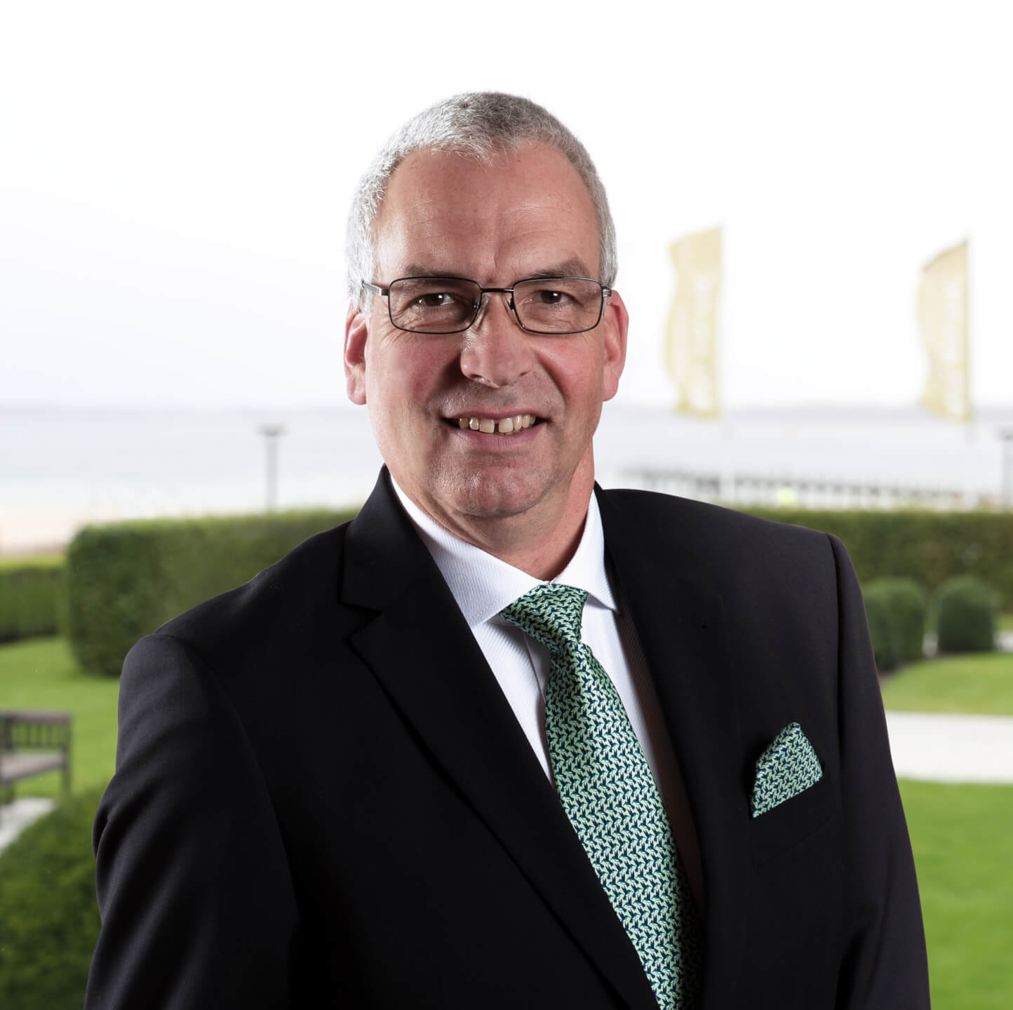 Hans-Peter Buerschaper - Restaurantleiter A man in a suit with glasses smiles, with the garden and sea view of the ATLANTIC Grand Hotel Travemünde in the background.