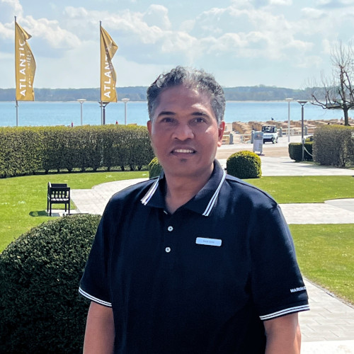 Made Soma from the ATLANTIC Grand Hotel Travemünde A hotel employee stands smiling in the garden of the ATLANTIC Grand Hotel Travemünde with the sea and flags in the background.