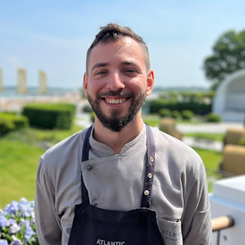 A smiling chef outdoors in front of the ATLANTIC Grand Hotel Travemünde with garden and beach in the background.