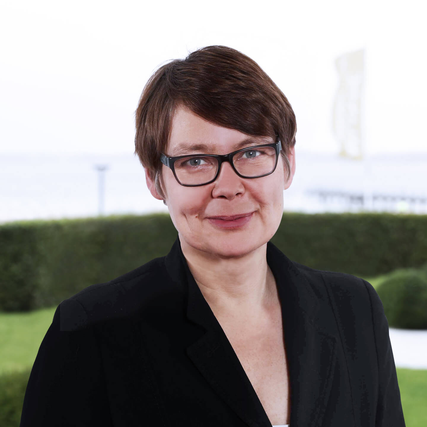 Housekeeper – Nicole Heitmann A smiling person with glasses in front of a green hedge and a view of the sea in the background.