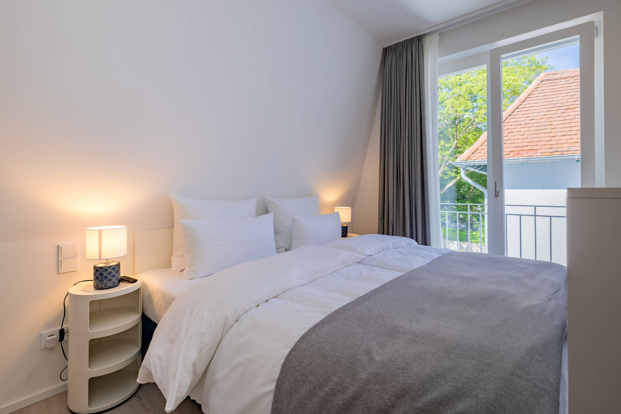 Helles Hotelzimmer mit Doppelbett, weißen Bettwaren, Nachttischlampen und Blick auf grüne Bäume durch große Fenster.