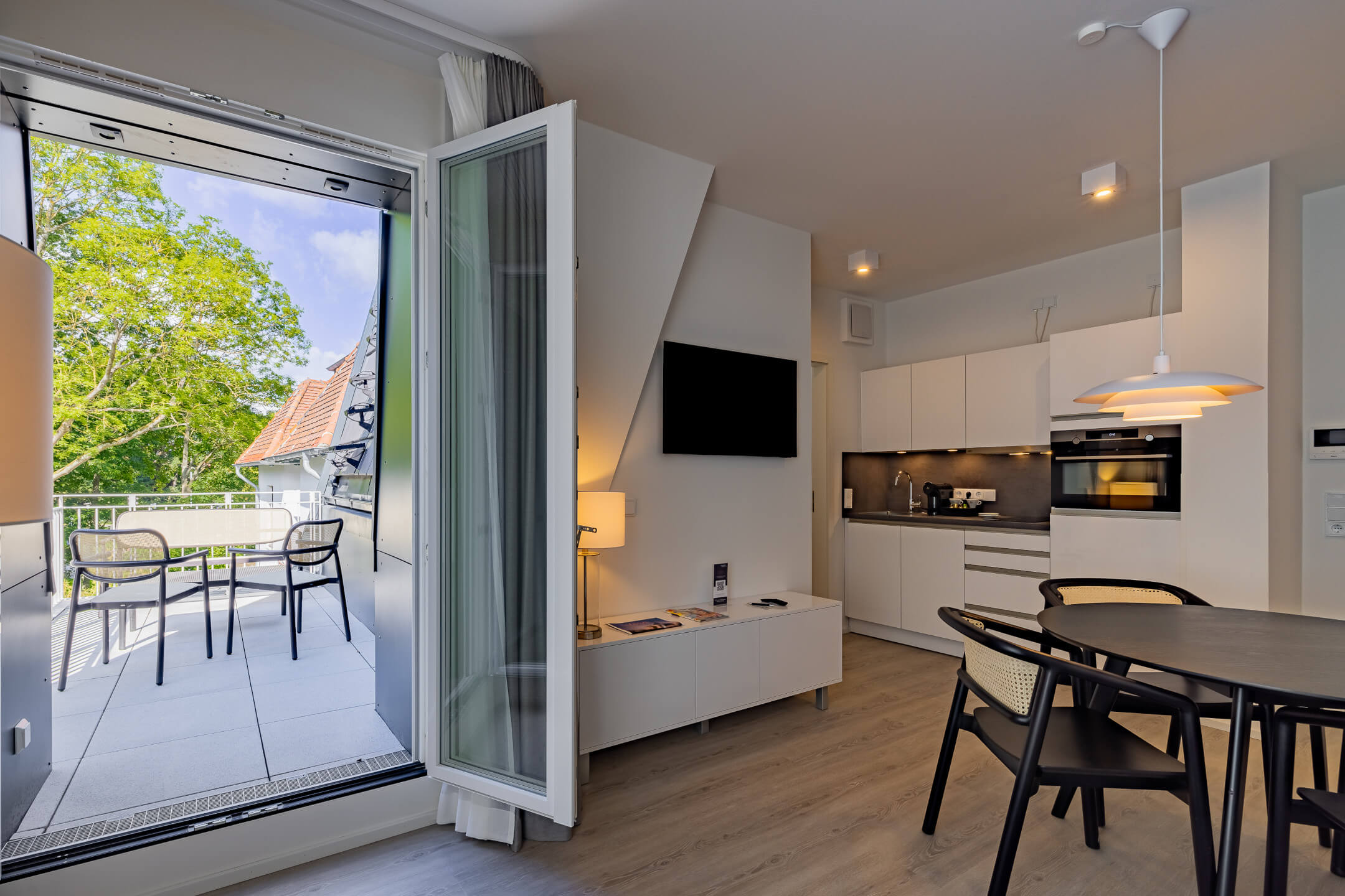 Bright hotel room with kitchenette, dining table and access to a balcony with seating area and view of green trees.