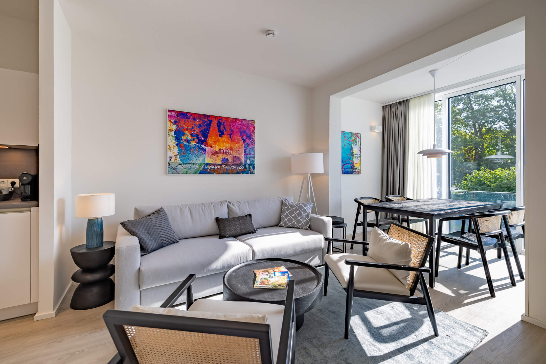 Modernes Hotelzimmer mit grauem Sofa, buntem Kunstwerk, Esstisch und großen Fenstern mit Blick ins Grüne.