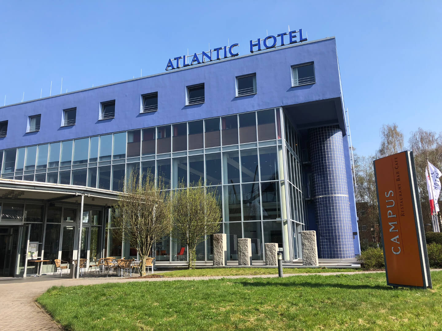 ATLANTIC Hotel Universum in Bremen, moderne Glasfassade, blauer Schriftzug, grüner Rasen, CAMPUS Restaurant Schild.
