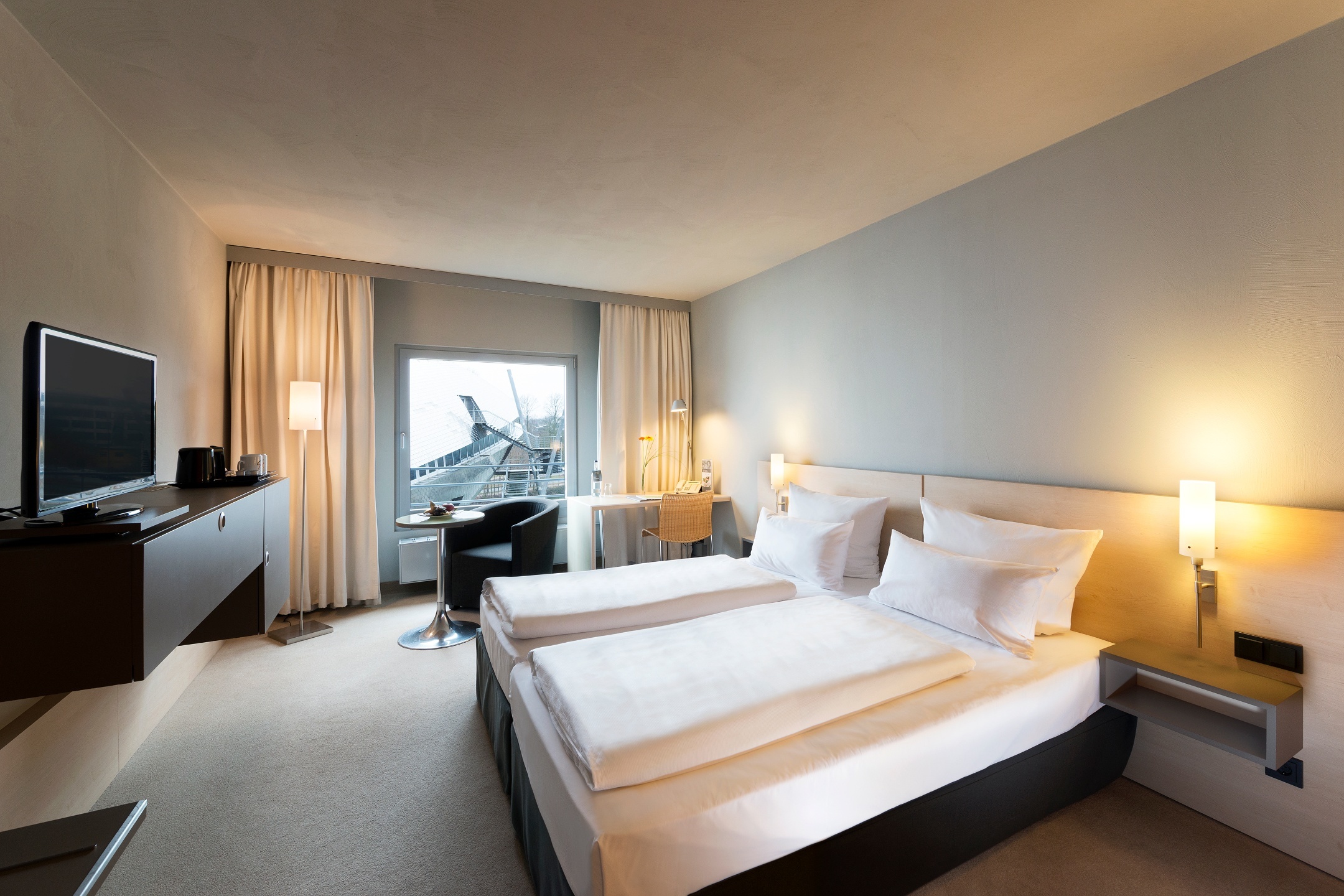 Bright hotel room with double bed, desk, TV and view of modern architecture through large window.
