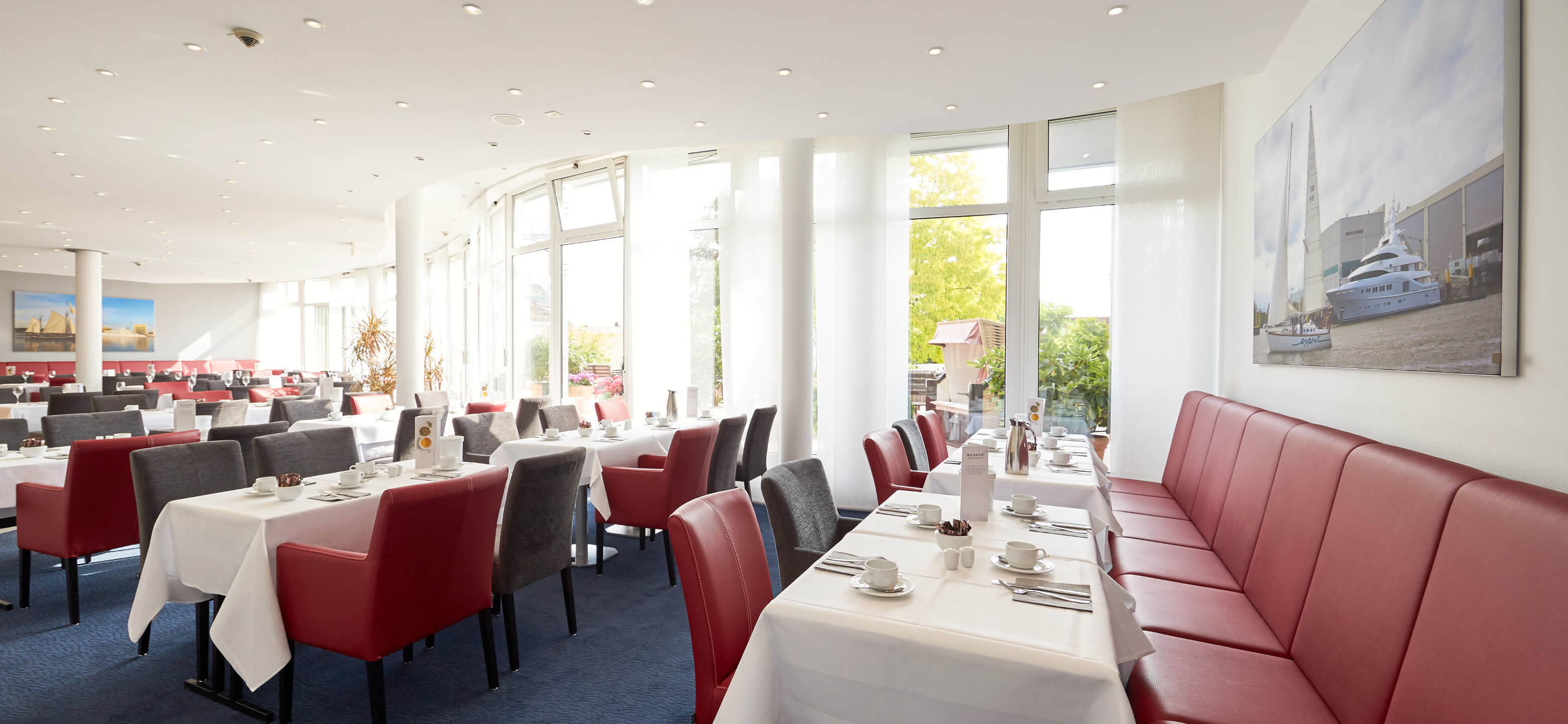 Helles Hotelrestaurant mit roten und grauen Stühlen, weiß gedeckten Tischen und großen Fenstern.