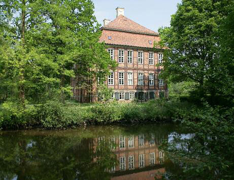 Historisches Backsteingebäude am Wasser, umgeben von grünen Bäumen, spiegelt sich im ruhigen Fluss.