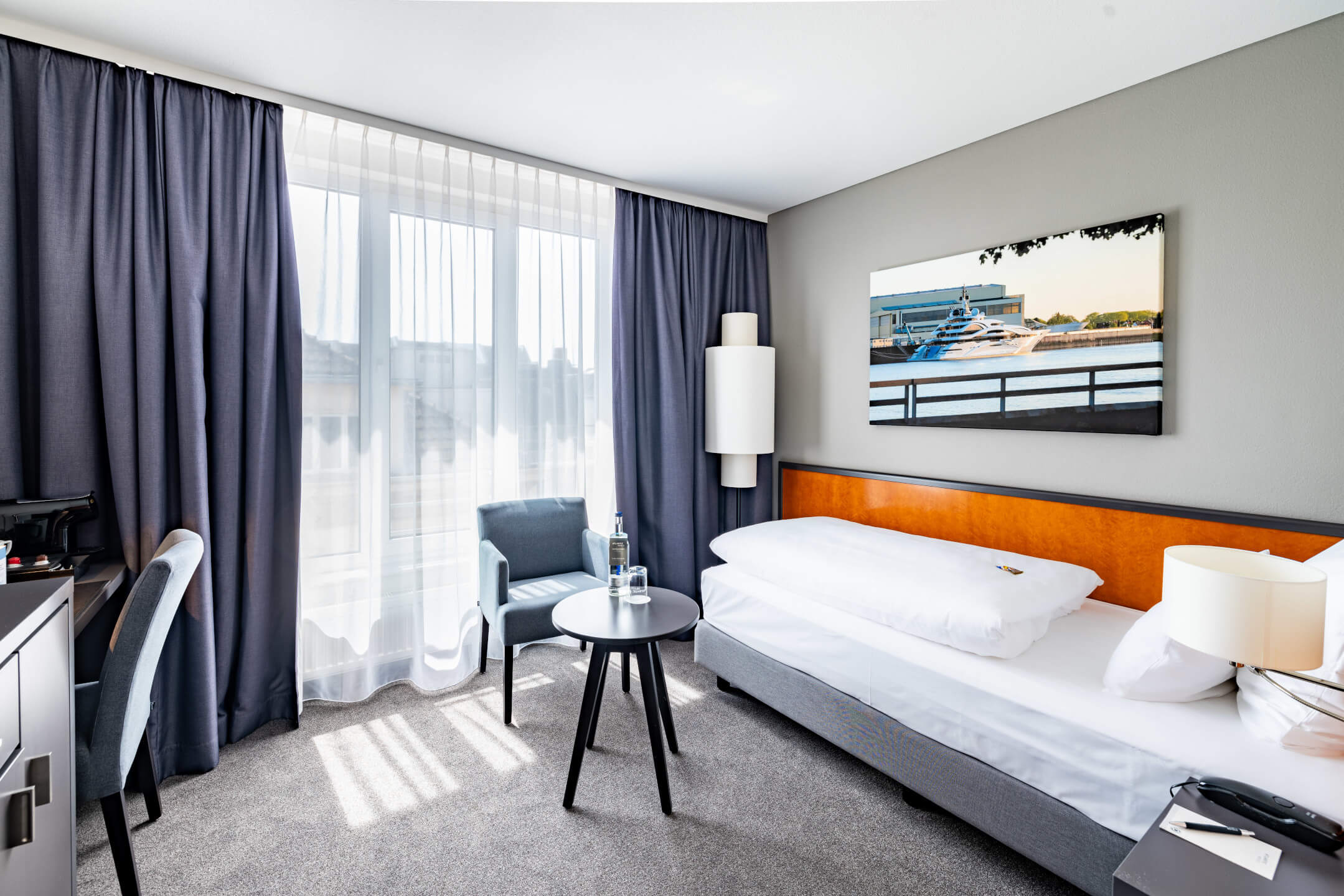 Helles Hotelzimmer mit Einzelbett, Schreibtisch, Sessel, rundem Tisch und großem Fenster im ATLANTIC Hotel Vegesack.