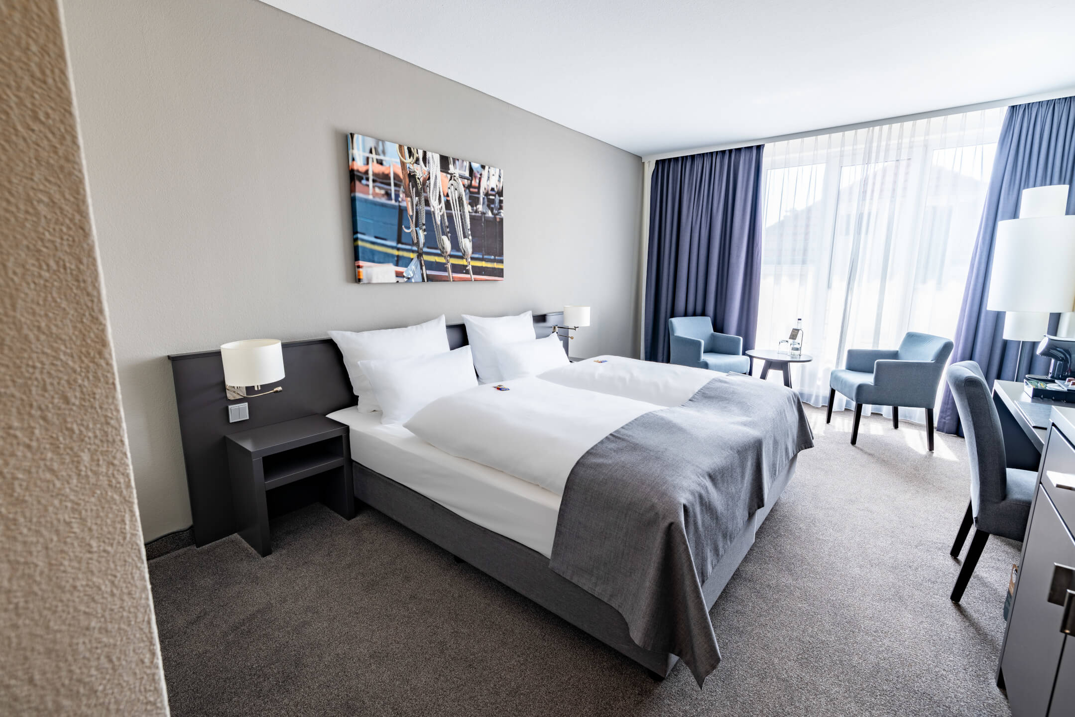 Helles Hotelzimmer mit Doppelbett, Sitzbereich, Schreibtisch und großem Fenster mit Vorhängen im ATLANTIC Hotel Vegesack.