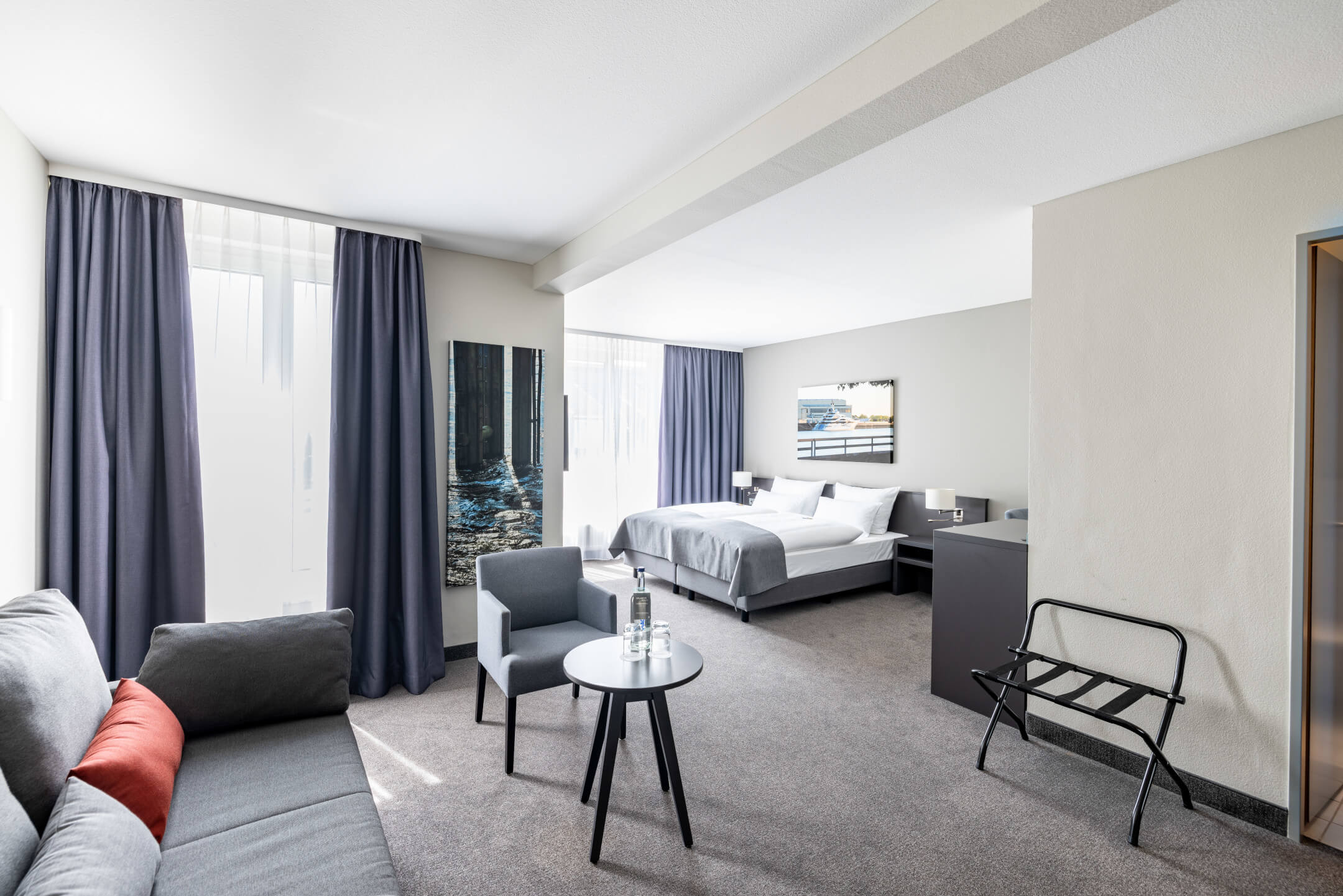 Helles Hotelzimmer mit Doppelbett, Sofa, Tisch, Stuhl und großen Fenstern mit blauen Vorhängen.