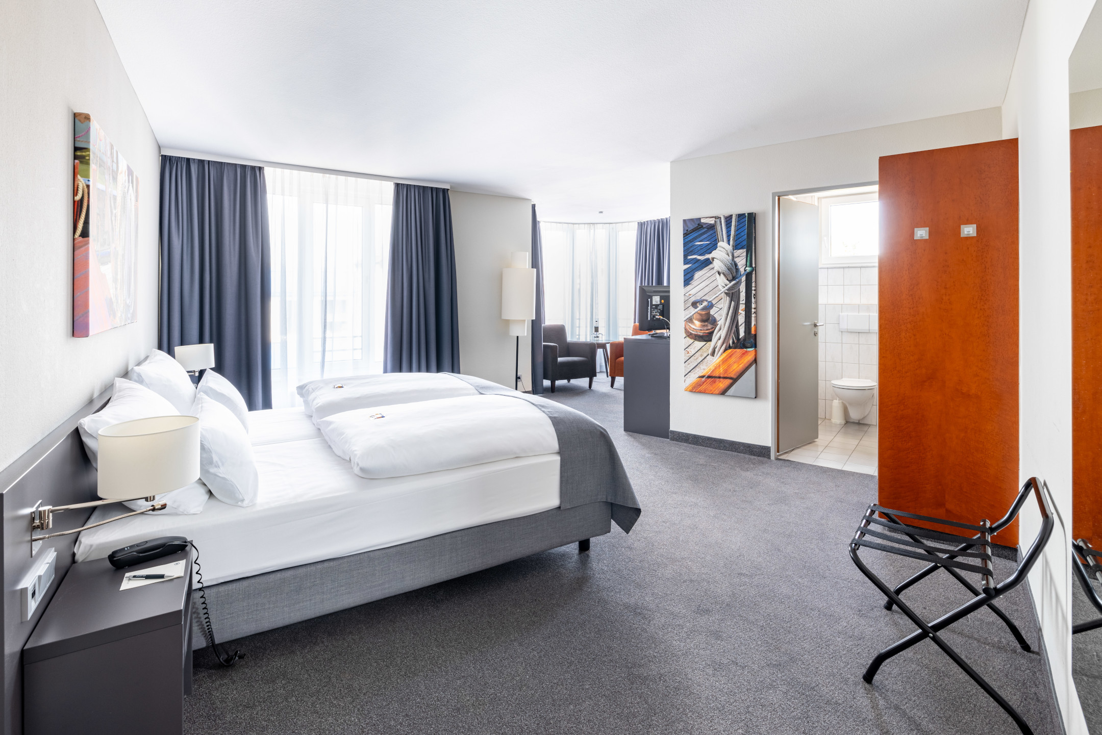 Helles Hotelzimmer mit Doppelbett, Sitzbereich, Kunstwerken und Zugang zu einem Badezimmer im ATLANTIC Hotel Vegesack.