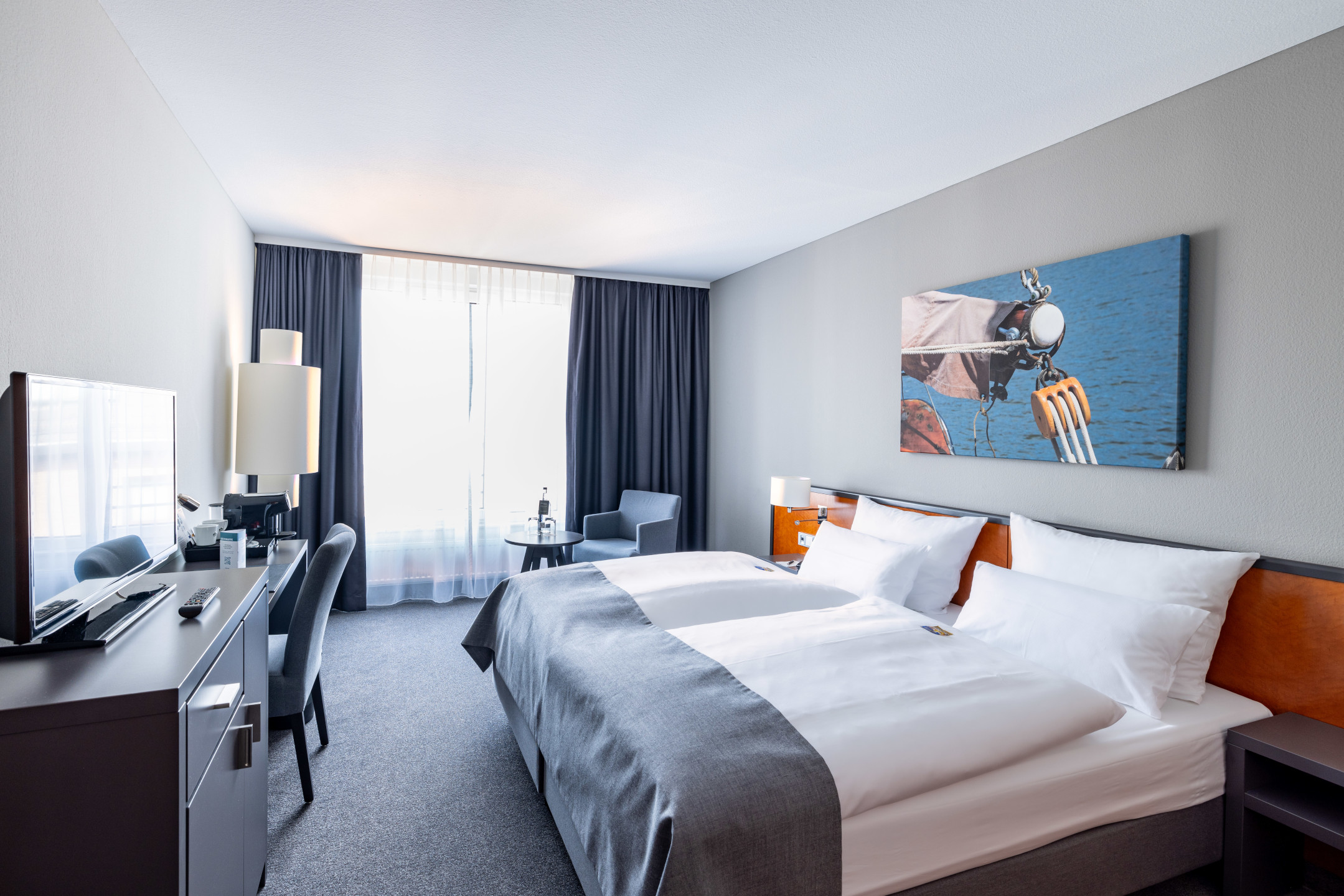 Helles Hotelzimmer mit Doppelbett, modernem Dekor, Schreibtisch, Sessel und großem Fenster im ATLANTIC Hotel Vegesack.