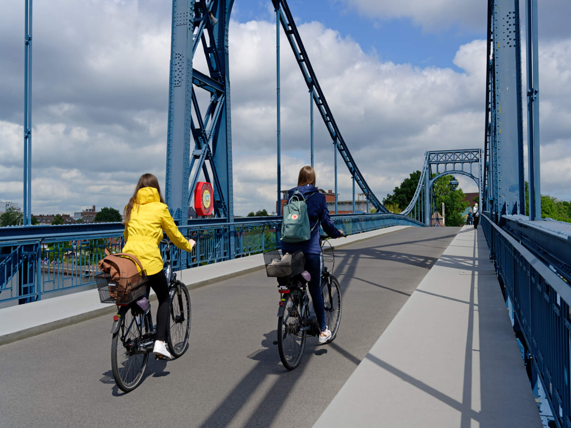 Zwei Personen radeln über eine blaue Stahlbrücke unter bewölktem Himmel.