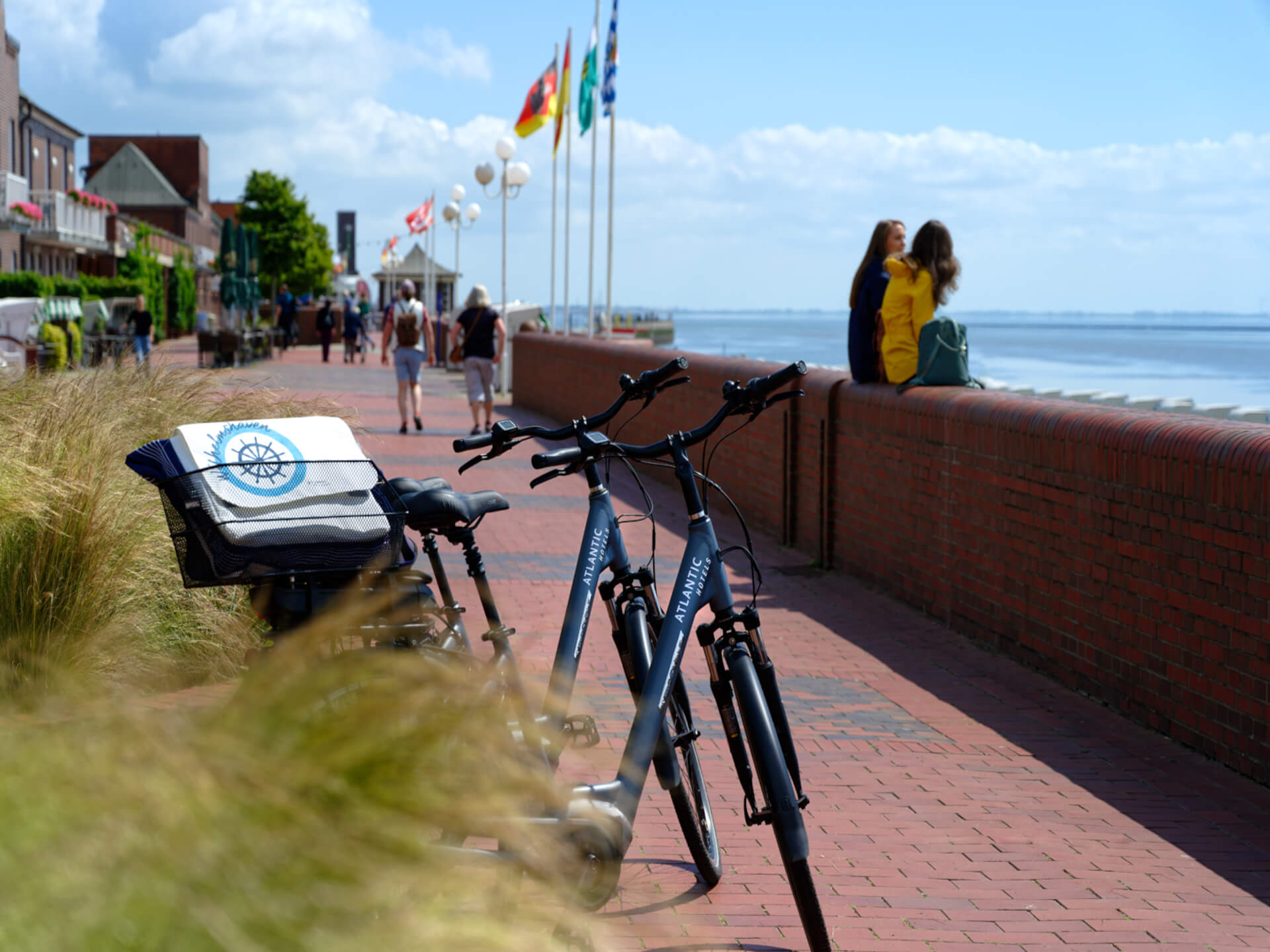 Fahrräder an einer Uferpromenade mit Blick aufs Meer, Menschen spazieren, Flaggen wehen im Wind.