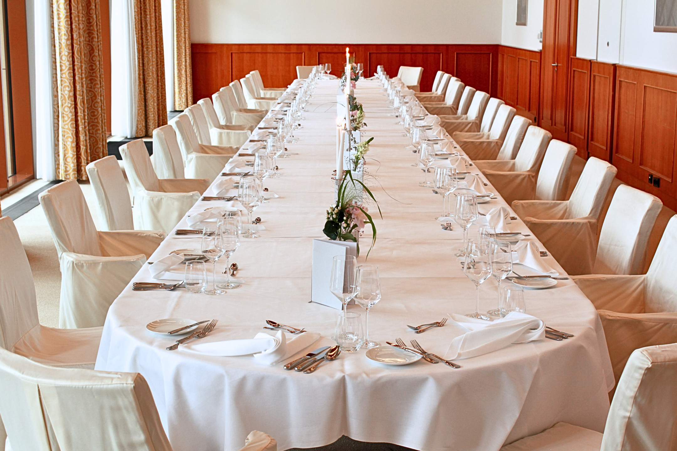 An oblong table laid for a festive occasion, with a white tablecloth, wine glasses, napkins, cutlery and chairs with fabric covers.