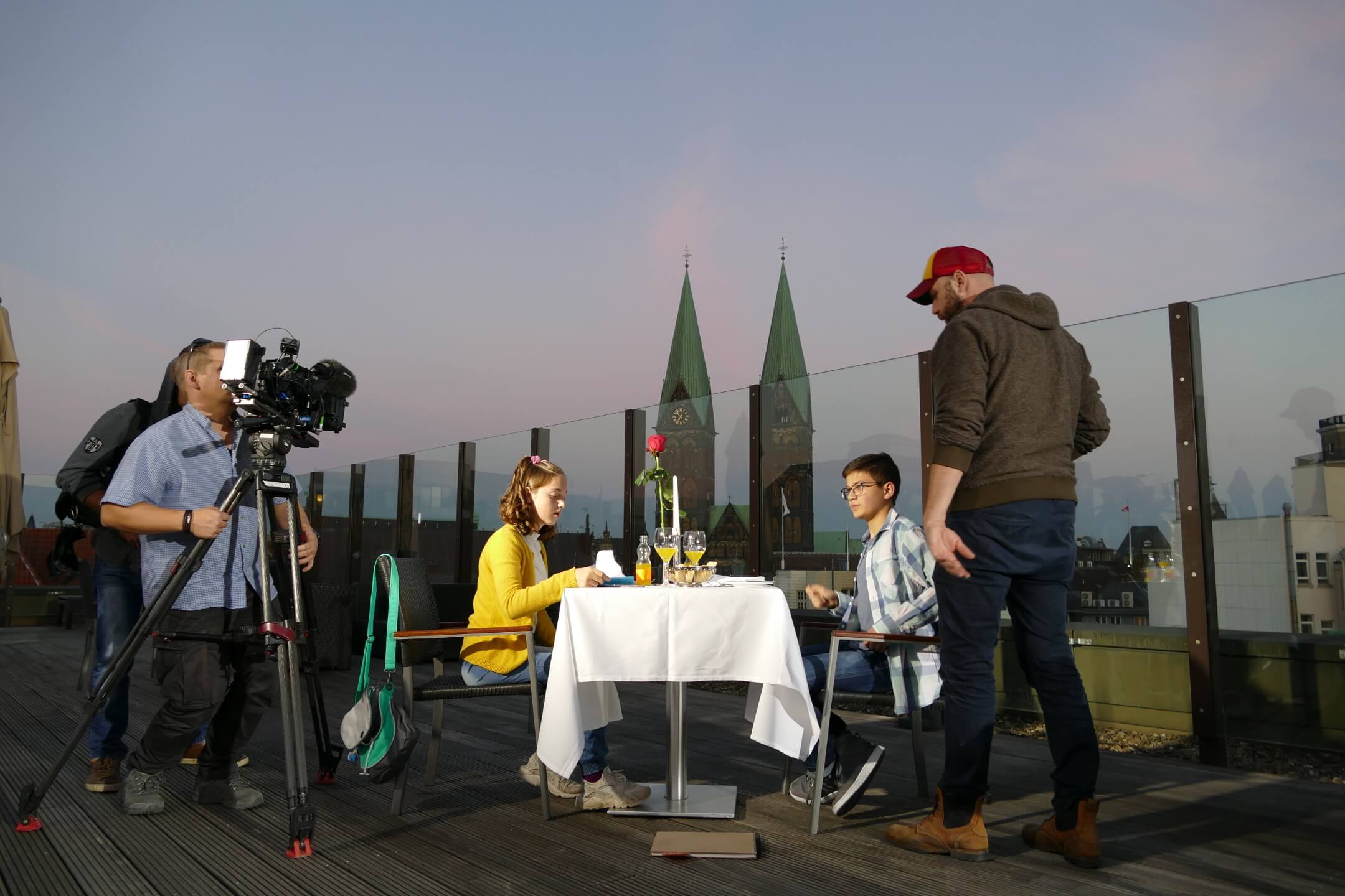 Web-Serie der ATLANTIC Hotels "Stille Post" Paar beim Abendessen auf einer Hotel-Dachterrasse, umgeben von Kamerateam, mit Blick auf Kirchtürme im Hintergrund.