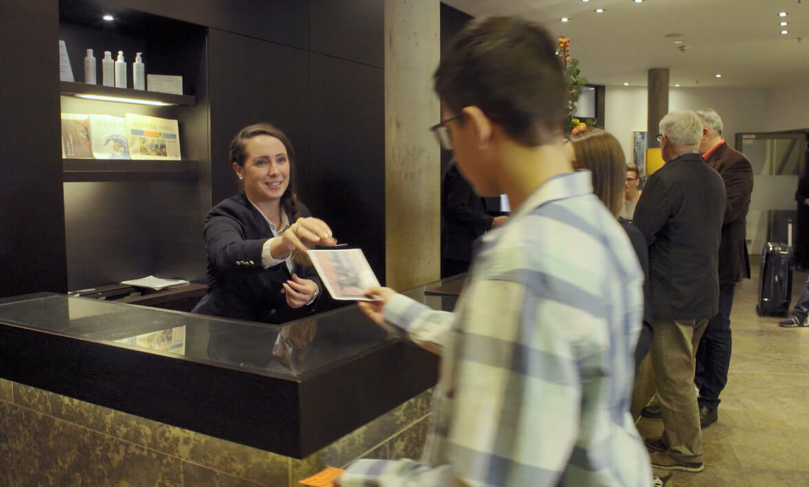 Rezeptionistin übergibt Gast ein Dokument, während andere Gäste im Hintergrund warten.