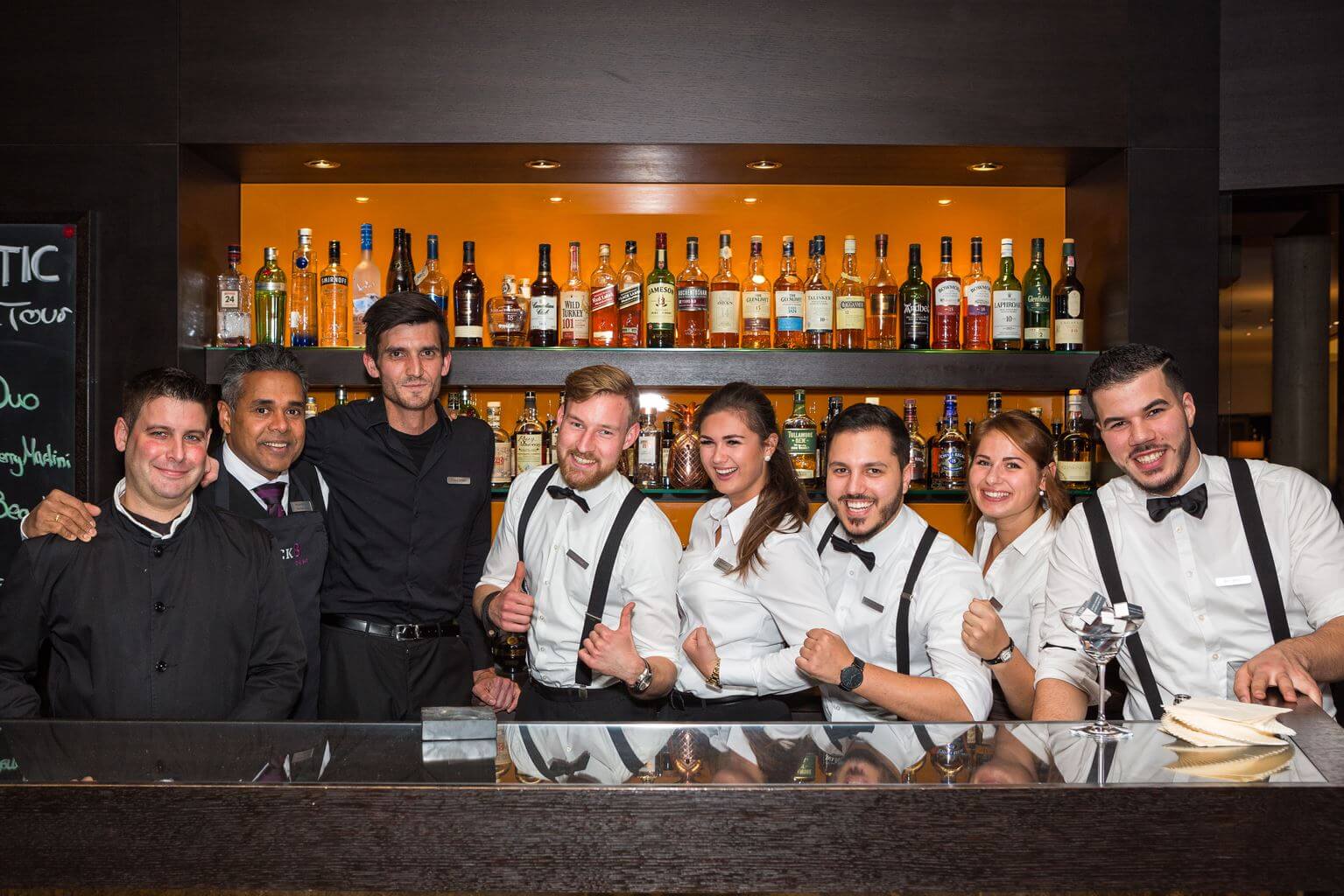 Barevent ATLANTIC Grand Hotel Bremen Bar team poses smiling in front of a well-stocked bar, all in elegant uniforms.