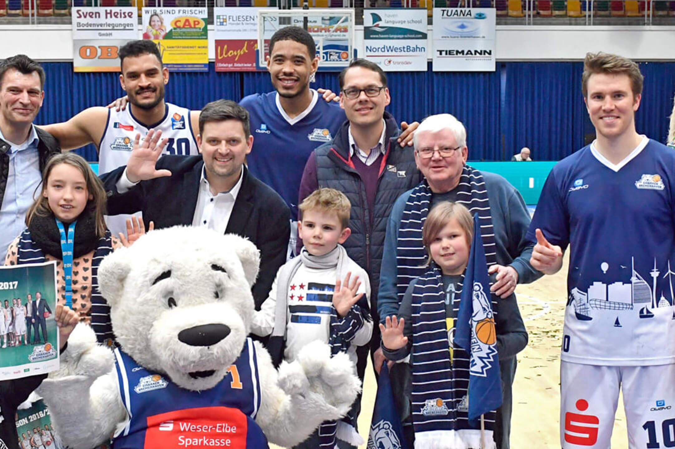 Group of people with basketball players and a mascot in a sports arena, all smiling and waving.