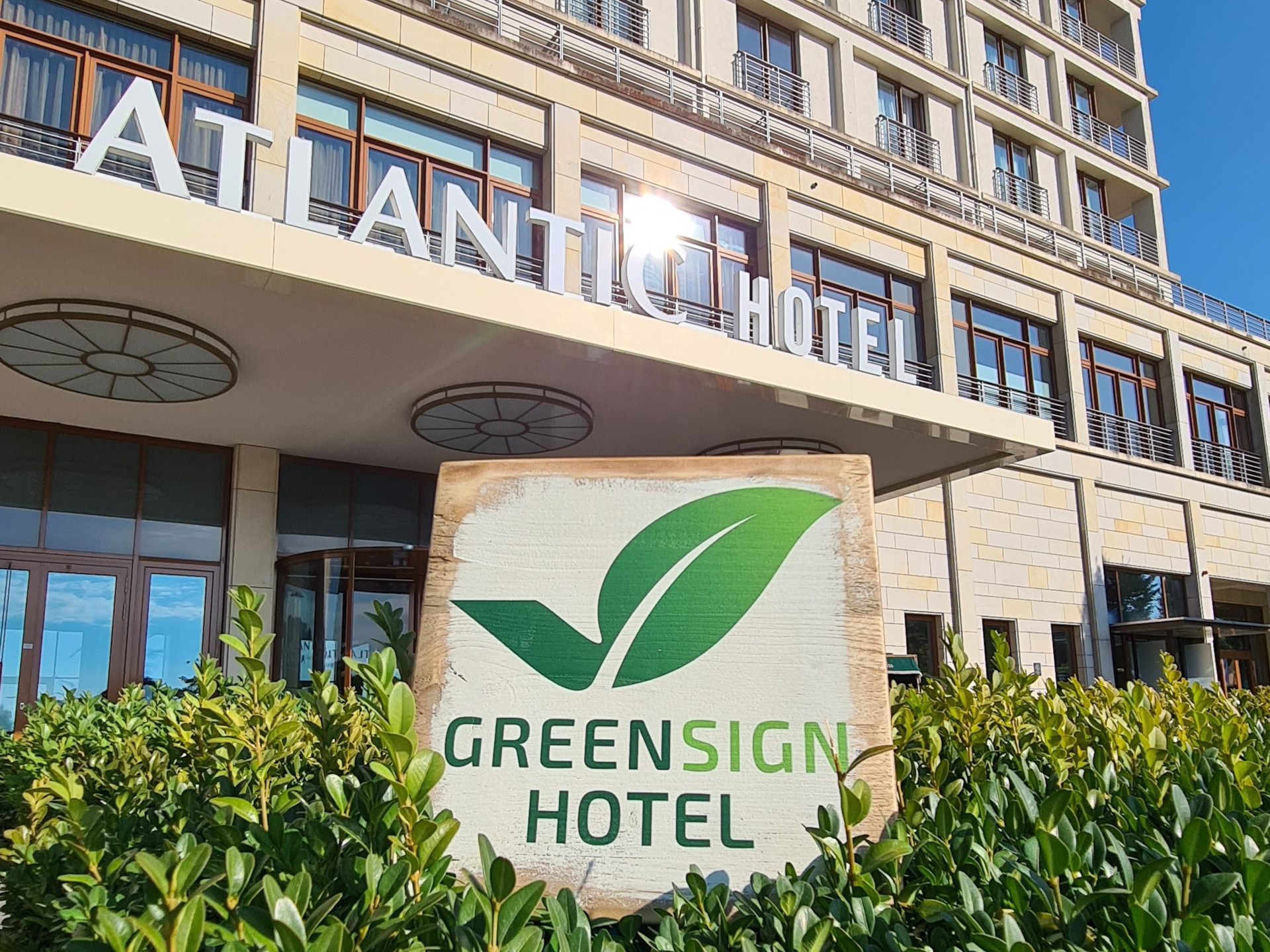 GreenSign Zertifikat | ATLANTIC Hotel Wilhelmshaven Fassade eines modernen Hotels mit "Greensign Hotel"-Schild und grünen Pflanzen im Vordergrund.