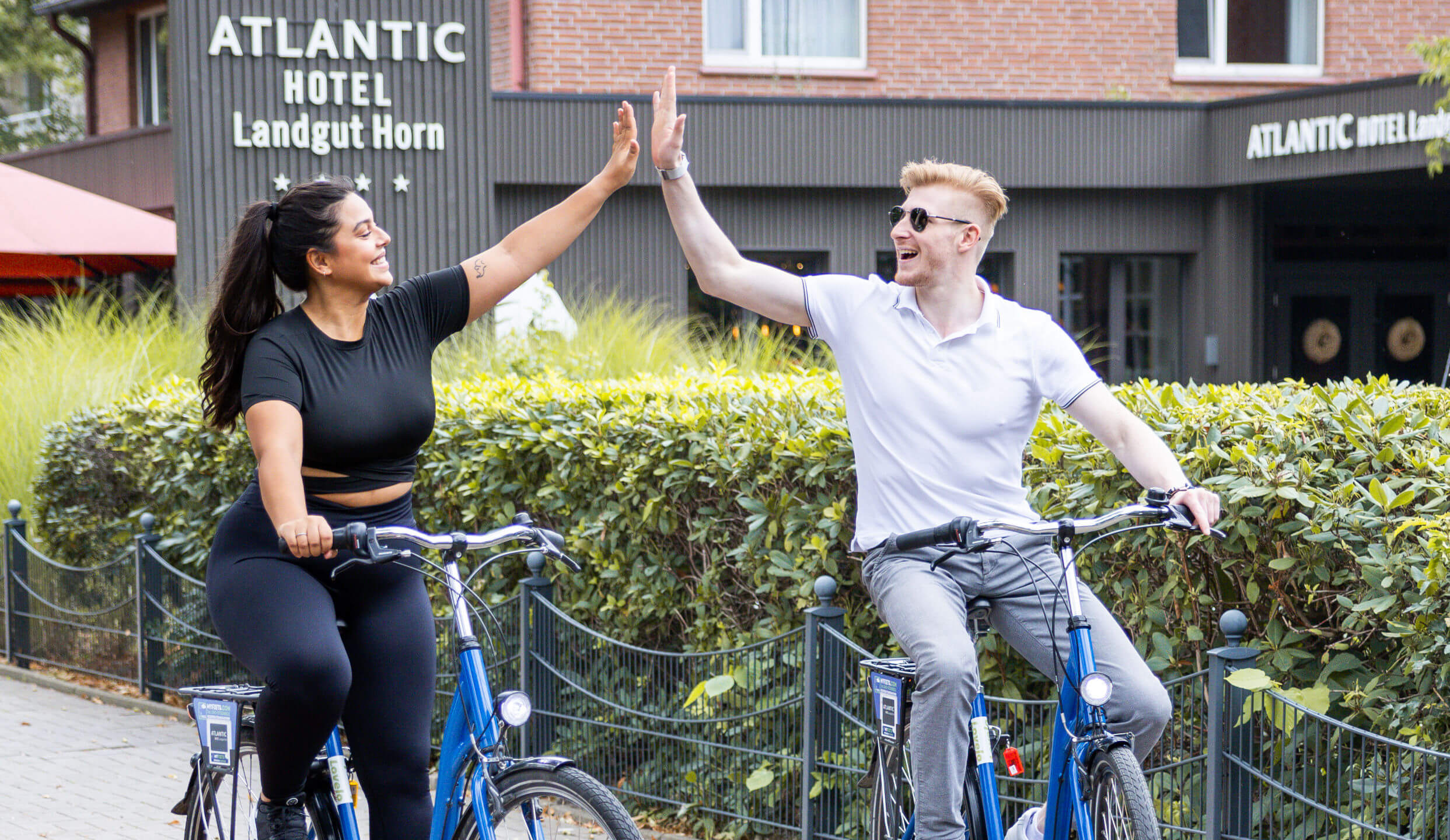 Zwei Personen auf Fahrrädern geben sich vor dem ATLANTIC Hotel Landgut Horn fröhlich ein High-Five.