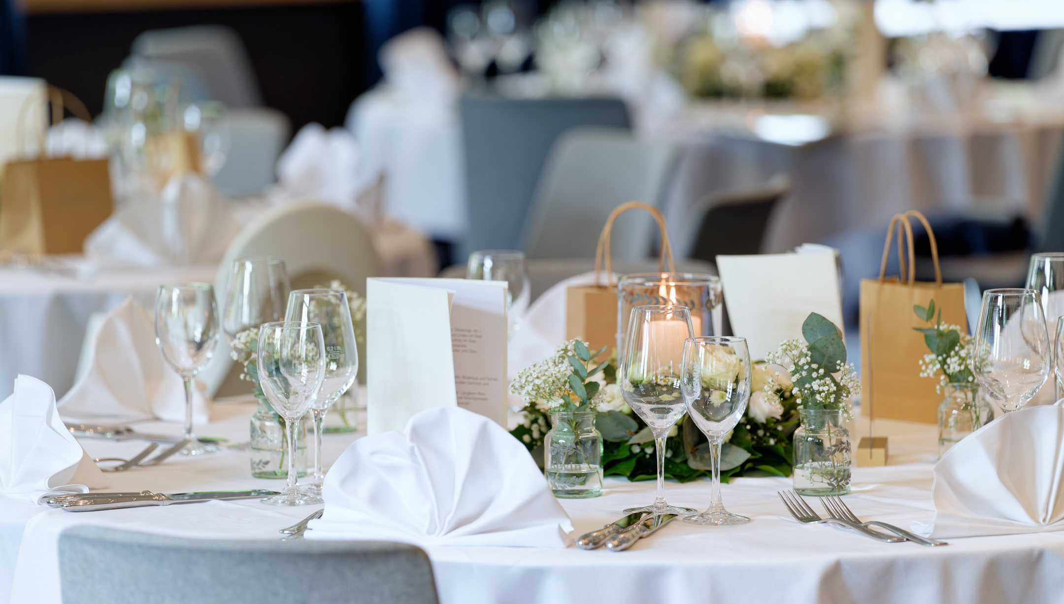Ein eingedeckter Tisch bei einer Hochzeit mit weißer Tischdecke, gefächerten Servietten, Schleierkraut und weißen Rosen.