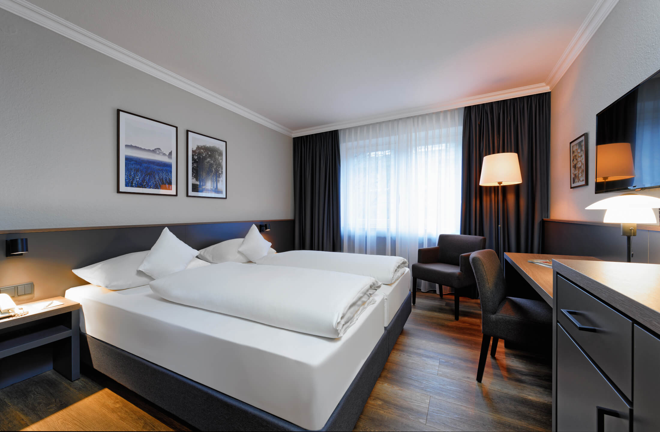 Modernes Hotelzimmer mit zwei Einzelbetten, Schreibtisch, Sessel, Stehlampe und Bildern an der Wand.