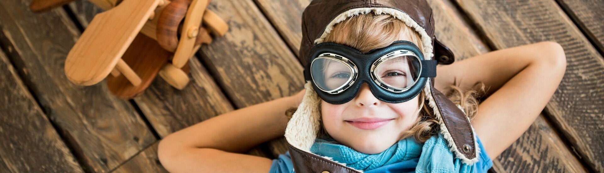Kind im ATLANTIC Hotel Airport in Bremen Kind mit Fliegermütze und Brille liegt lächelnd auf Holzboden neben Holzflugzeug.
