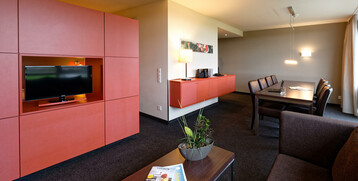 Modern hotel room with red wardrobe, flat-screen TV, dining table, plants and cozy seating area at the ATLANTIC Hotel Bremen.