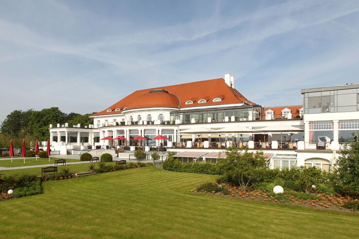 Elegant hotel with a red roof, large windows and terraces, surrounded by well-tended gardens with flowers and lawns.