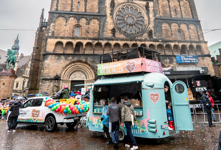 Ein bunter Foodtruck mit Ballons steht vor einer historischen Kirche, umgeben von Menschen bei Regenwetter.