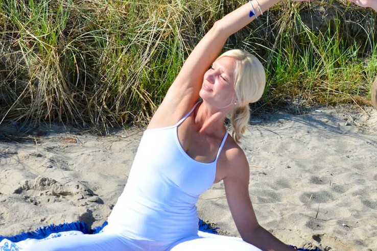 Frau in weißer Yoga-Kleidung macht Yoga am Sandstrand vor Dünen auf blau-weißer Decke.
