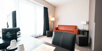 Helles Hotelzimmer mit rotem Sofa, Schreibtisch, TV, zwei Kaffeetassen und großen Fenstern mit Vorhängen.