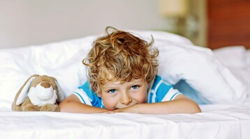 Child lies smiling on a hotel bed with a cuddly toy, surrounded by white pillows. Photo: © Irina Schmidt - Fotolia.com