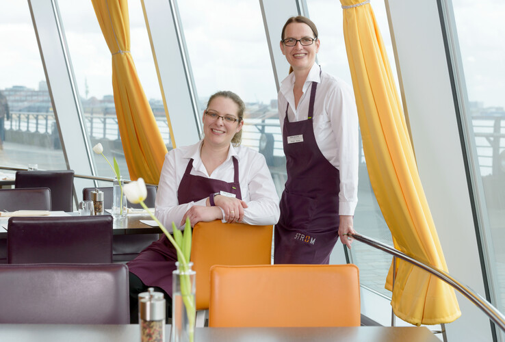 Two employees in the restaurant of the ATLANTIC Hotel Sail City, with a view of large windows and yellow curtains.