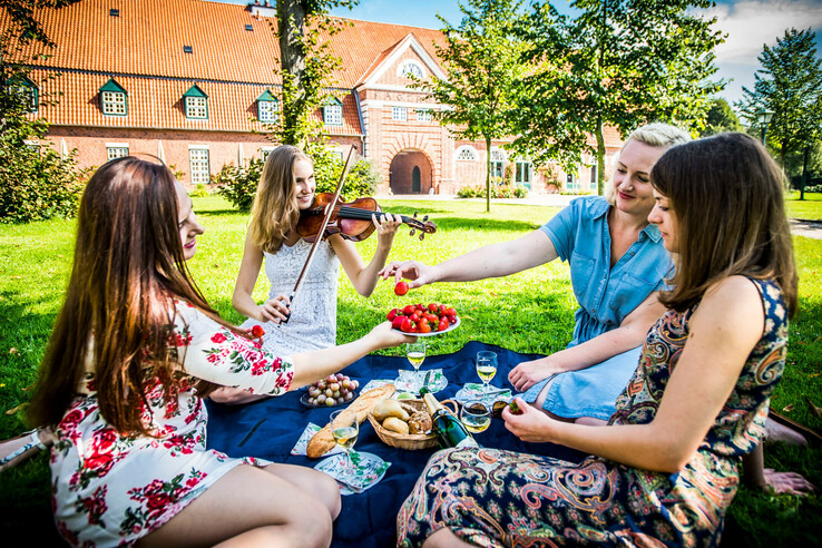 Vier Frauen picknicken auf einer Wiese vor einem Backsteingebäude, eine spielt Geige.