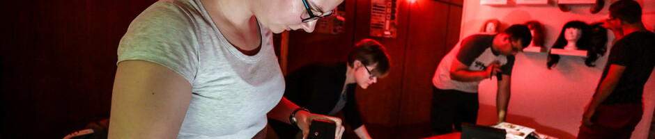 Personen lösen Rätsel in einem rot beleuchteten Escape Room mit Perücken an der Wand.