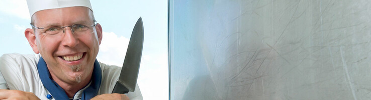 A chef in uniform smiles and holds two knives in front of a window in the ATLANTIC Hotel Sail City, Bremerhaven.