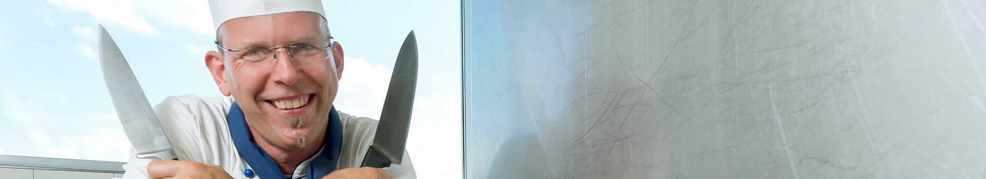 A chef in uniform smiles and holds two knives in front of a window in the ATLANTIC Hotel Sail City, Bremerhaven.