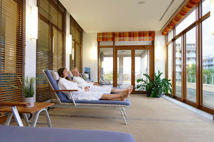 Two people relax in bathrobes on loungers in a bright room with large windows and plants.