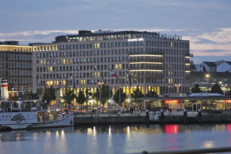 Hotel building on the waterfront at dusk, illuminated, with flags and restaurant in the foreground.