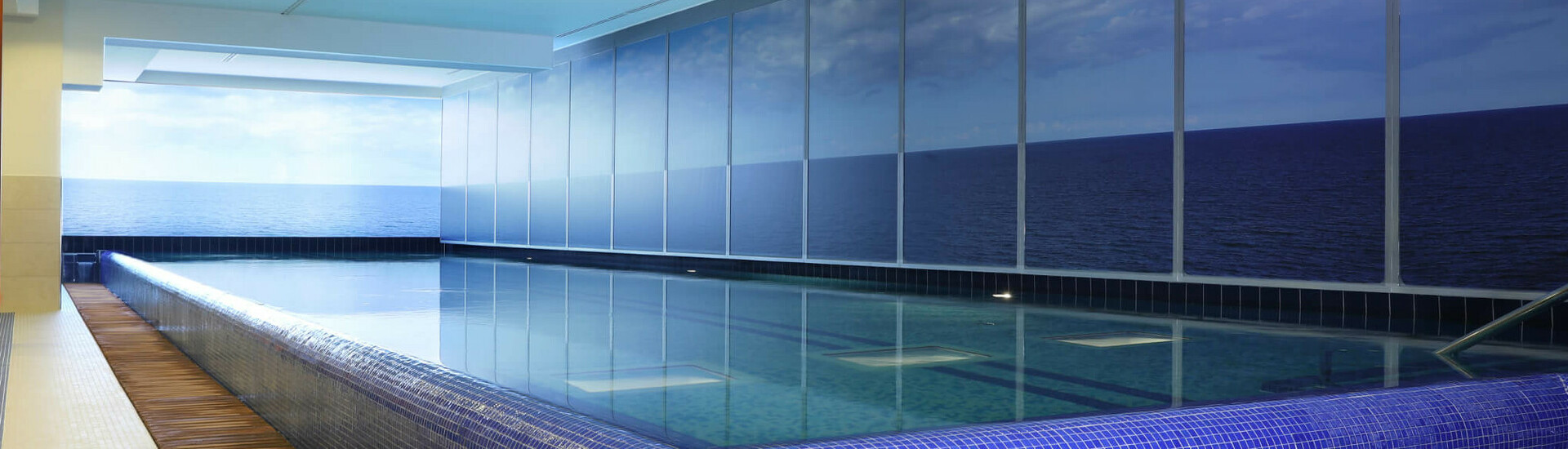 Schwimmbad ATLANTIC Grand Hotel Travemünde Innenpool mit blauem Mosaikrand und Meerblick durch große Fenster im ATLANTIC Grand Hotel Travemünde.