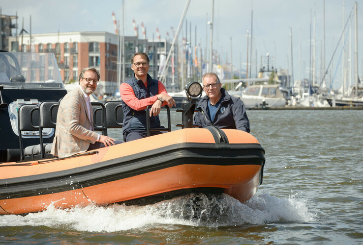 Drei Personen auf einem orangefarbenen Schnellboot im Hafen von Bremerhaven, mit Segelbooten im Hintergrund.