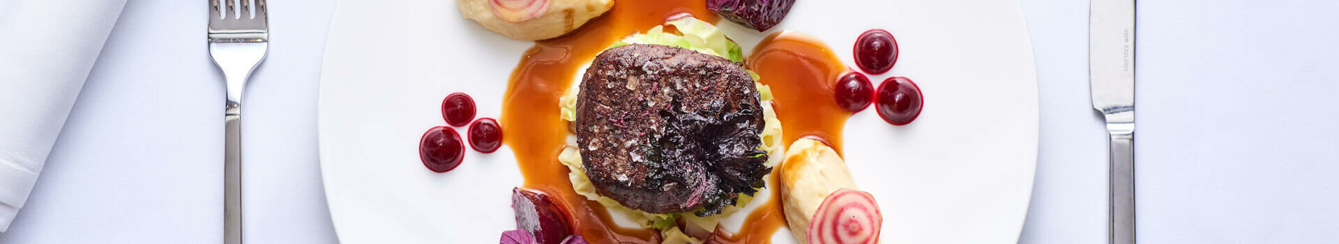 Gourmetgericht auf weißem Teller: Steak mit Sauce, Beilagen und Dekoration, auf weißer Tischdecke angerichtet.