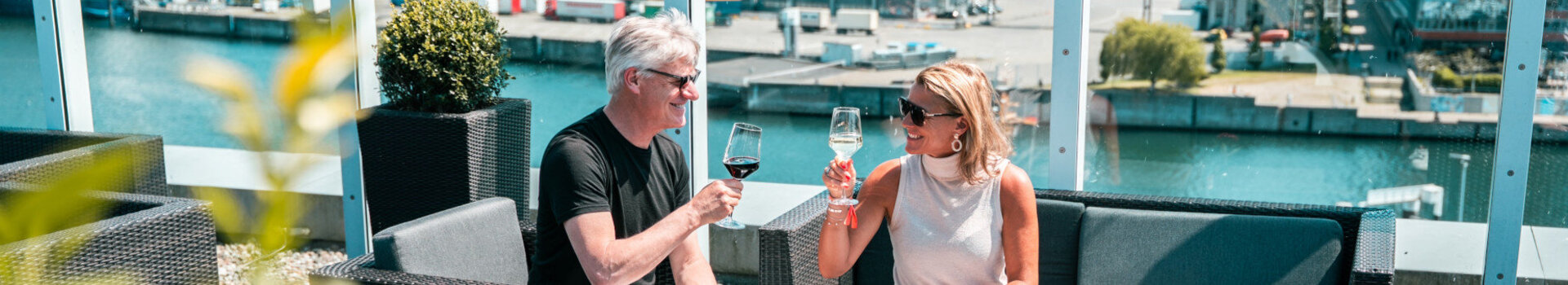 A man and a woman clink glasses, laughing, whilst sitting on the sunny roof terrace.