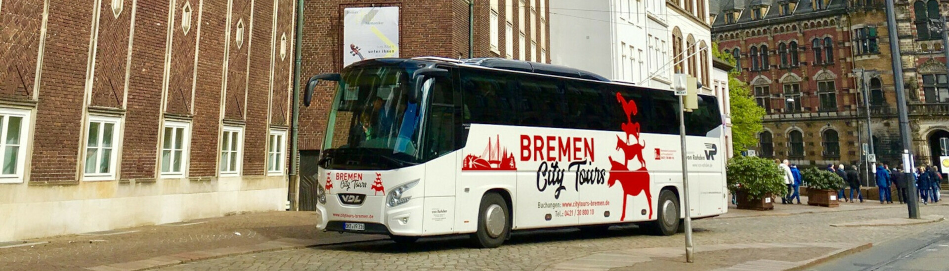 Bremer City Tours l Rahmenprogramm ATLANTIC Hotel Galopprennbahn Ein weißer Tourbus mit Bremen-Stadtmusikanten-Logo parkt vor historischen Gebäuden in Bremen.