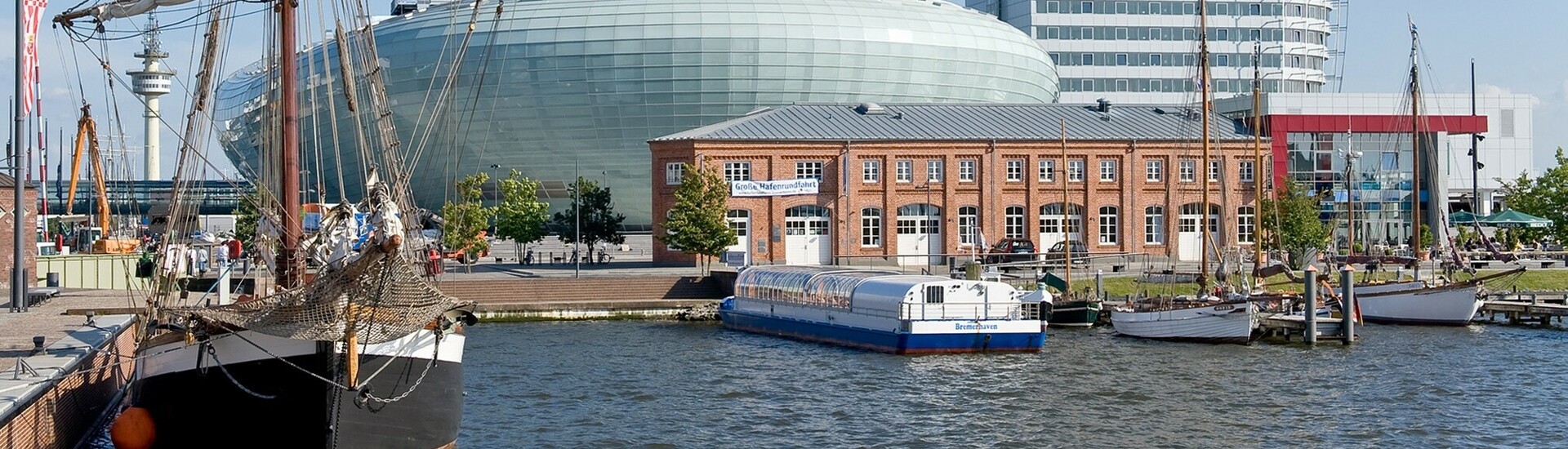 Hafenansicht mit Segelschiff, modernem Glasgebäude und historischem Backsteingebäude bei klarem Himmel.