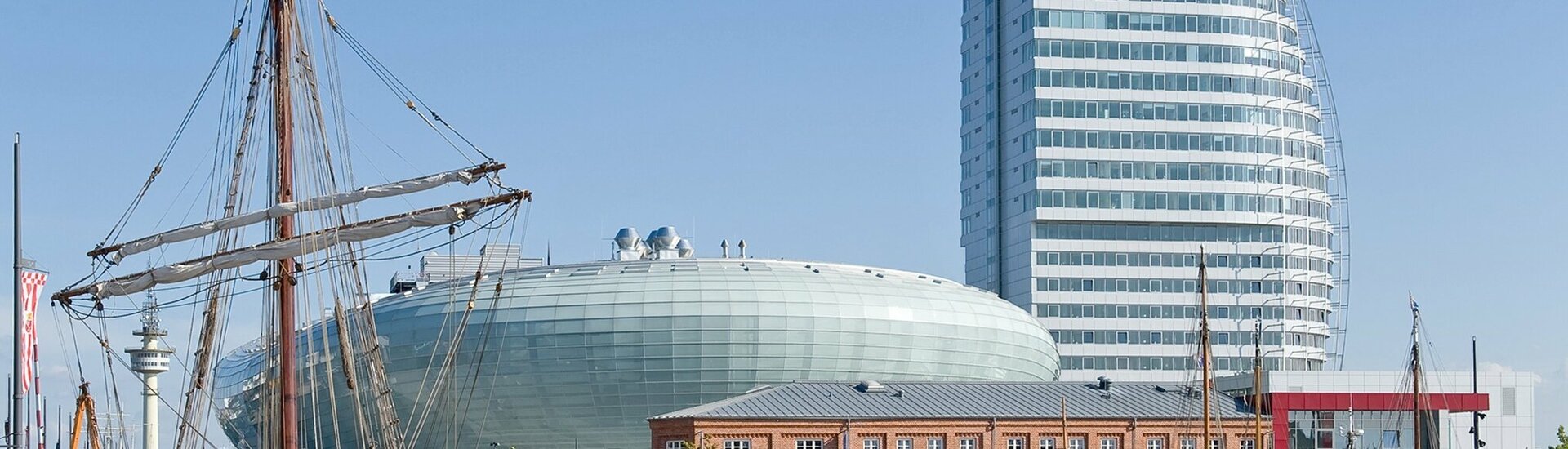 Harbor view with sailing ship, modern glass building and historic brick building under a clear sky.